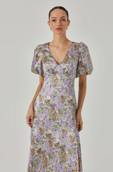 A woman is wearing a long dress with a floral pattern in shades of purple, green, orange, and cream. The dress has short, puffed sleeves with slight gathers at the shoulders, a V-neckline, and a fitted waist. The fabric appears to have a textured or slightly shiny surface.